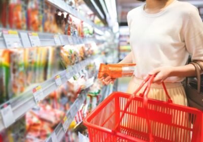 Customer with a red basket taking a packet from a supermarket shelf lined with printed price labels, an example of store labeling