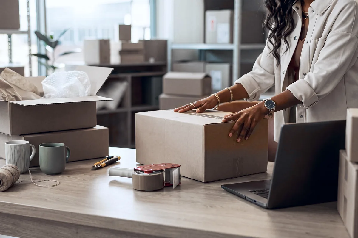 Online seller sealing a cardboard shipping box at a desk beside a laptop and packing supplies